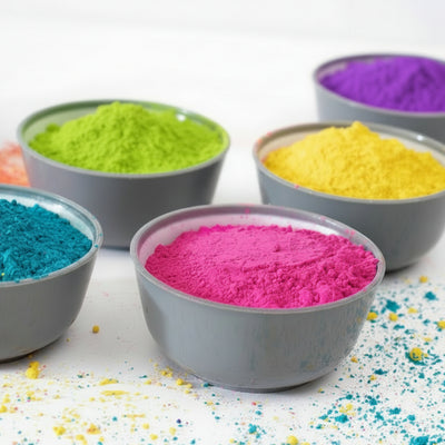 Five bags of bulk premium colour powder in different colors (blue, pink, green, yellow, purple) displayed on a table with some color powder scattered around.